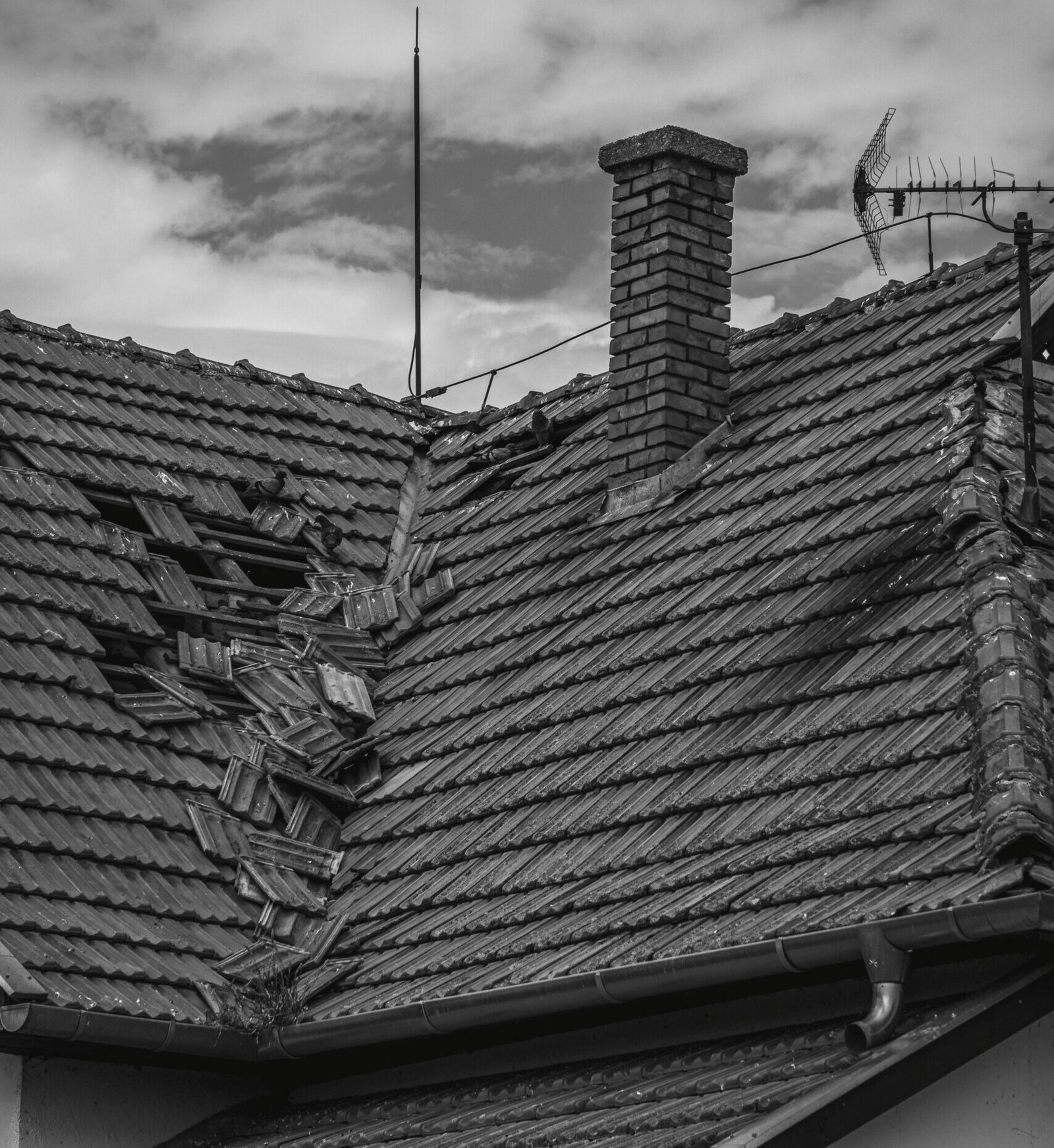 A grayscale photo of a broken roof with a chimney and antenna under a cloudy sky.