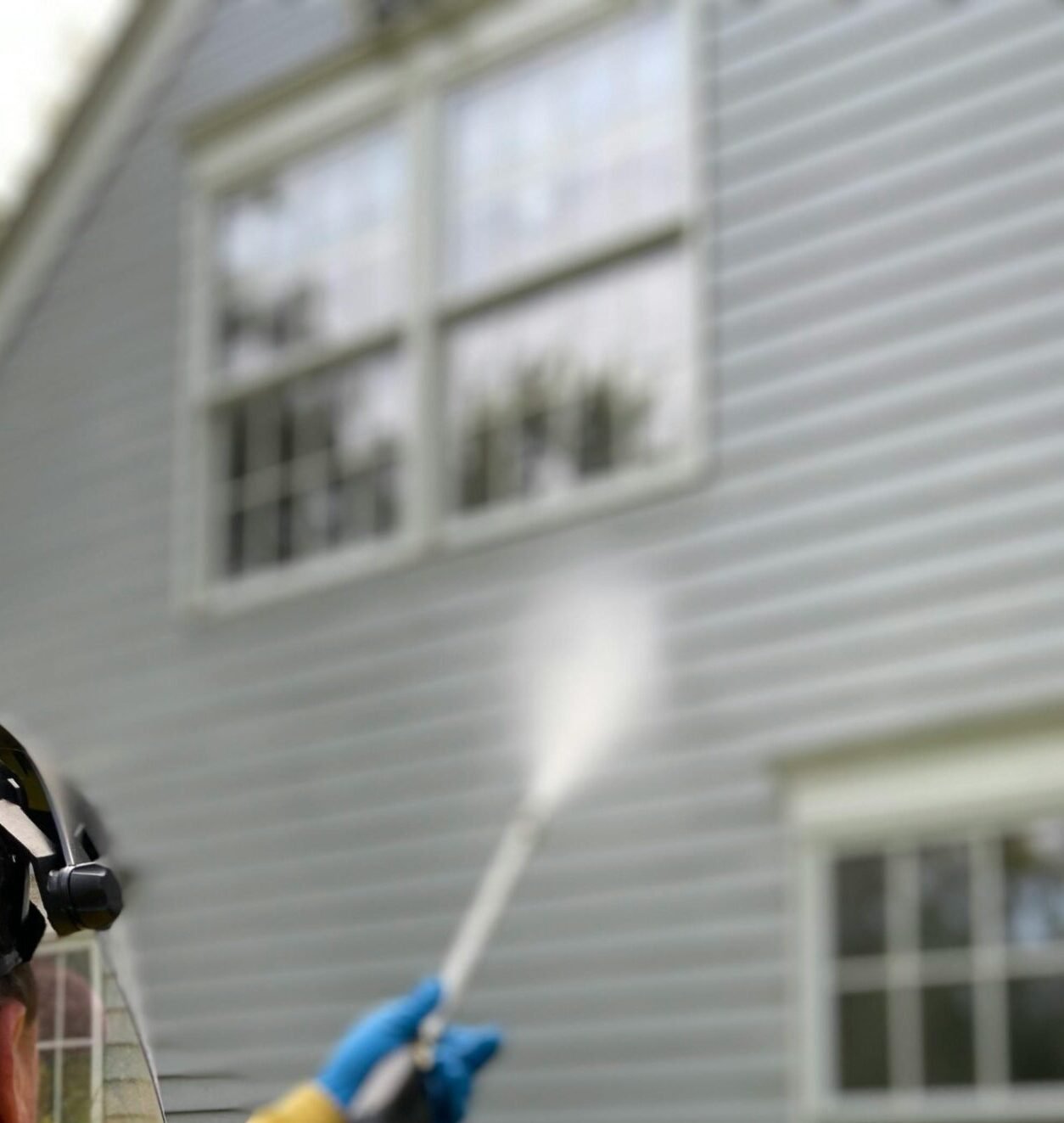 Person cleaning house siding with a pressure washer, wearing protective gear in a yellow raincoat.